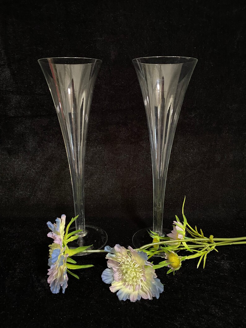May include: Two clear glass champagne flutes with a fluted design. The flutes are on a black surface with blue and green flowers.