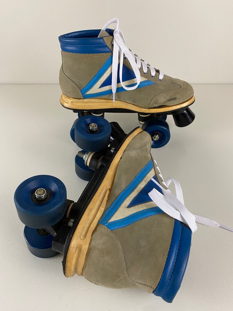 May include: A pair of vintage roller skates with blue and white accents. The skates are made of suede and have a high-top design. The wheels are blue and have a black center.