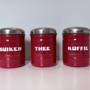 Op de afbeelding: Drie bijpassende rode blikken met zilveren deksels. De blikken hebben witte tekst met de tekst "SUKER", "THEE" en "KOFFIE". De blikken zijn cilindrisch en lijken van metaal te zijn.