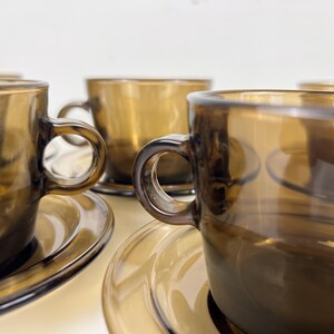 K&ouml;nnte beinhalten: Set aus vier Teetassen und Untertassen aus bernsteinfarbenem Glas. Die Tassen haben abgerundete Griffe und die Untertassen einen leicht erh&ouml;hten Rand.