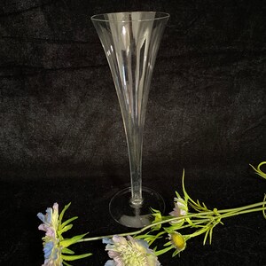 May include: A clear glass champagne flute with a tall, slender stem and a wide, flared bowl. The flute is sitting on a black surface with two blue and white flowers.