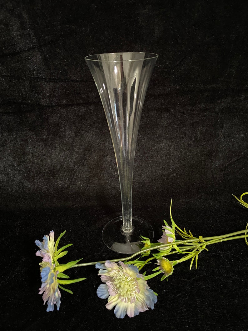 May include: A clear glass champagne flute with a tall, slender stem and a wide, flared bowl. The flute is sitting on a black surface with two blue and white flowers.