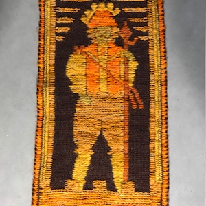 May include: A brown and orange woven wall hanging with a stylized figure of a person holding a staff. The figure is wearing a hat and has a long, flowing robe.