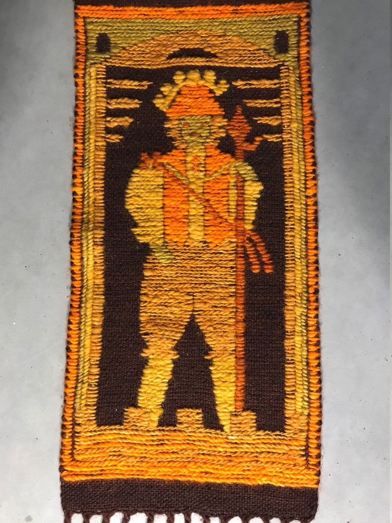 May include: A brown and orange woven wall hanging with a stylized figure of a person holding a staff. The figure is wearing a hat and has a long, flowing robe.