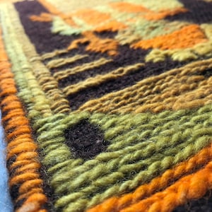 May include: A close-up of a woven wall hanging with a geometric pattern in shades of brown, orange, and green.