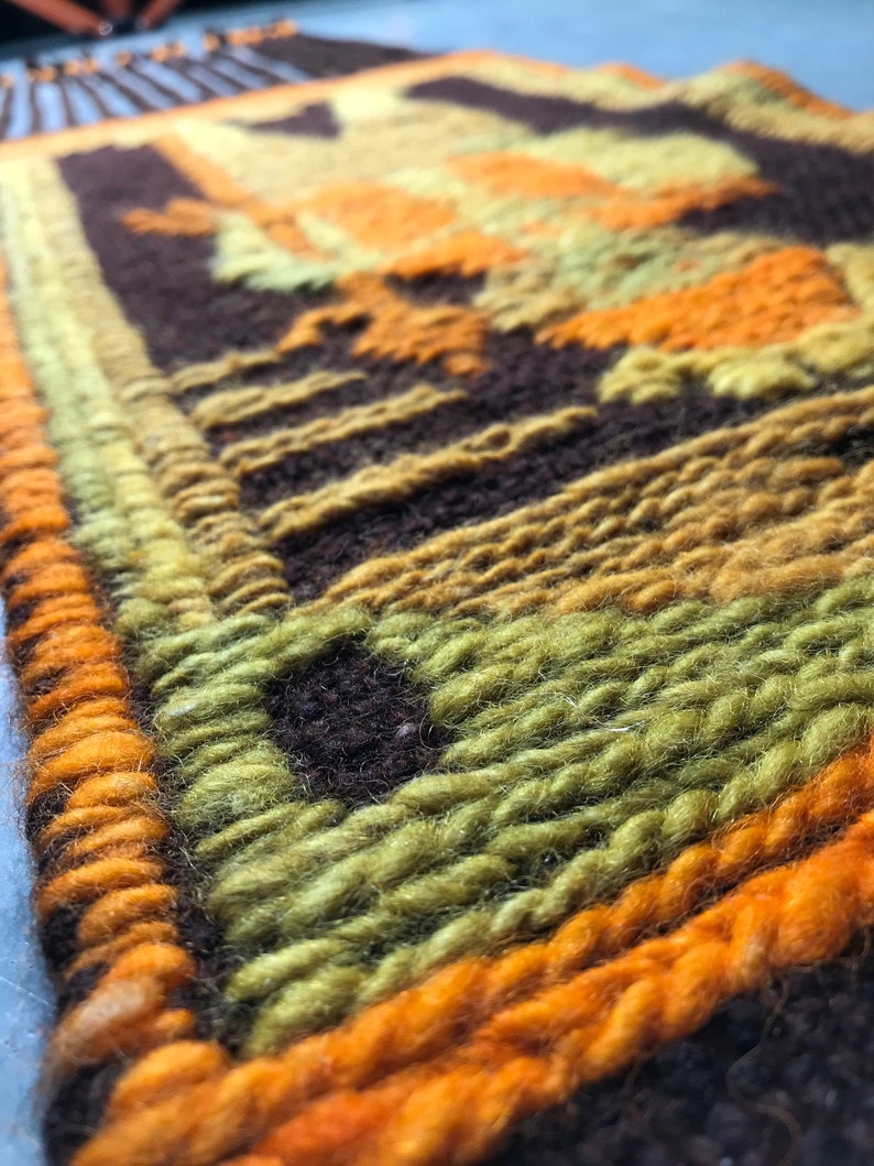 May include: A close-up of a woven wall hanging with a geometric pattern in shades of brown, orange, and green.