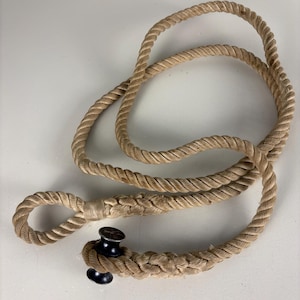 Puede incluir: Una cuerda beige con un lazo en un extremo y una cornamusa negra en el otro. La cuerda está enrollada y anudada, lo que sugiere un tema náutico o decorativo. La cornamusa es de metal negro.