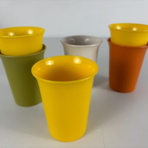 Op de afbeelding: Een verzameling kleurrijke plastic bekers in tinten geel, oranje, groen en gebroken wit. De bekers hebben een uitlopend ontwerp en zijn in paren gestapeld, waarbij de ene beker in de andere is genest. De bekers zijn gerangschikt op een lichtgrijze ondergrond.