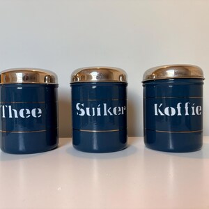 Op de afbeelding: Drie bijpassende donkerblauwe bussen met goudkleurige banden en deksels. Elke bus heeft een wit label met een ander woord: "Thee", "Suiker" en "Koffie". De bussen zijn in een rij op een wit oppervlak geplaatst.