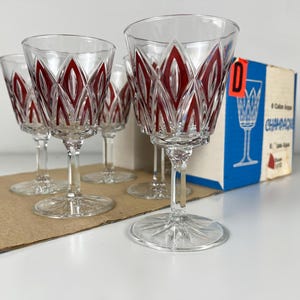 May include: Set of six clear glass champagne flutes with a red and white geometric pattern. The flutes are arranged on a brown surface with a box labelled "Champagne" in the background.