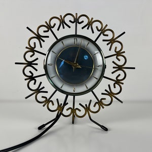 Op de afbeelding: Een vintage mid-century modern klok met een zwarte wijzerplaat en gouden accenten. De klok heeft een uniek ontwerp met een ronde lijst en een standaard met drie poten. De klok is aangesloten en klaar om de tijd aan te geven.
