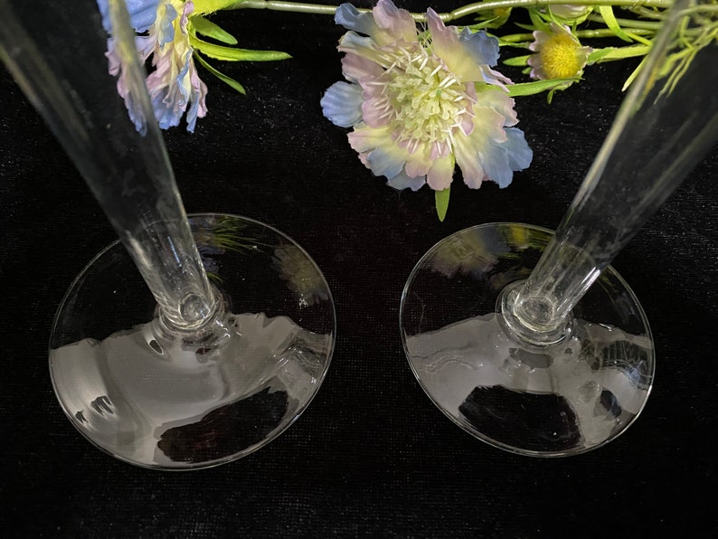 May include: Two clear glass candle holders with a wide base and a tall, slender stem. The candle holders are on a black surface.