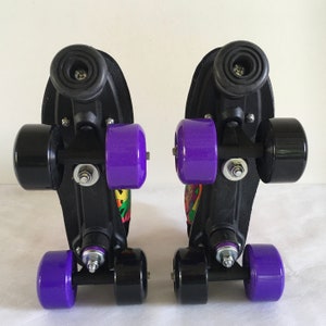 May include: Pair of black roller skates with purple wheels and colorful graphics on the sides. The skates have a black metal frame and a black plastic base.