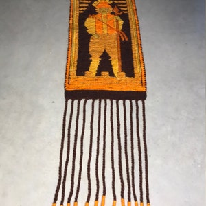 May include: A vintage woven wall hanging with a brown and orange geometric design of a person holding a staff. The hanging has a fringe of brown yarn with orange accents.