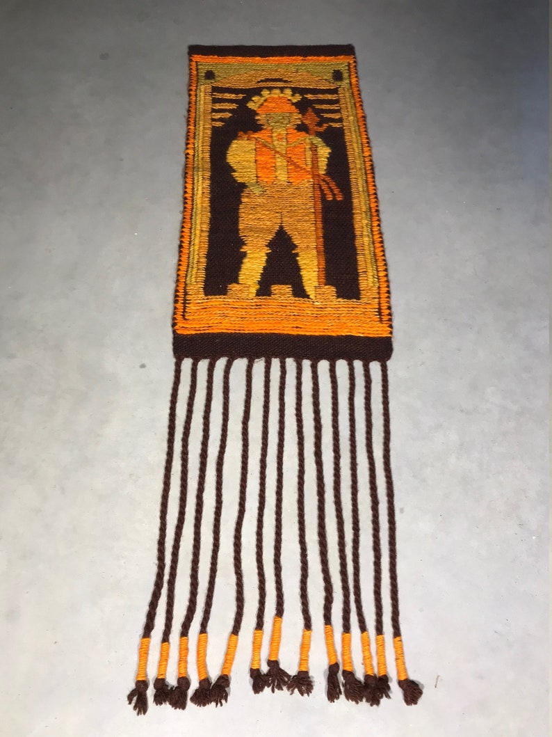 May include: A vintage woven wall hanging with a brown and orange geometric design of a person holding a staff. The hanging has a fringe of brown yarn with orange accents.