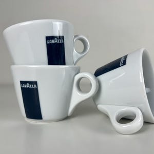 May include: Three white ceramic espresso cups with a navy blue rectangular logo that says "Lavazza". The cups have a unique handle design and are stacked on a white surface.