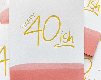May include: A white card with a pink watercolor wash at the bottom. The card says "Happy 40-ish" in gold lettering.