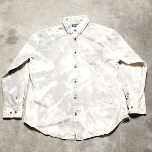 May include: A gray button-down shirt with a bleached, tie-dye effect. The long-sleeved shirt has a button-down collar, a chest pocket, and buttoned cuffs. The shirt is laid flat on a concrete surface.