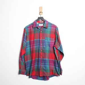 Puede incluir: Una camisa de cuadros de manga larga en rojo, verde azulado, morado y verde cuelga de una percha negra. La camisa tiene una parte delantera abotonada, un cuello y un bolsillo en el pecho. El estampado de cuadros es un diseño clásico.