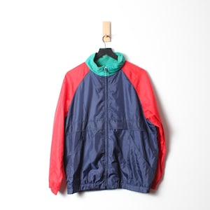 May include: A vintage windbreaker jacket in a color block design. The jacket features red sleeves, a navy blue body, and a teal green collar. It has a full zipper closure and is hanging on a wooden hook against a white background.