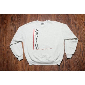 May include: A light gray heathered crewneck sweatshirt featuring a vertical red line next to the black text Aastrom and the smaller red text BIOSCIENCES, INC. printed on the front. The garment has long sleeves and a ribbed collar and cuffs.