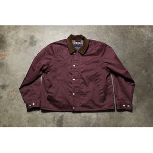 May include: A burgundy jacket with a brown corduroy collar and a plaid lining. The jacket has a button-up front, two front pockets, and buttoned cuffs. The jacket is made of a smooth, water-resistant material.
