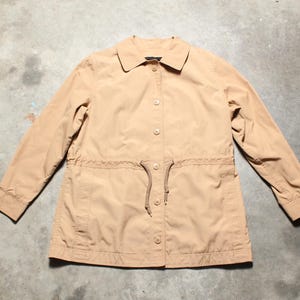 May include: A tan-colored jacket with a collared neckline and long sleeves. The jacket features a button-up front and a drawstring waist. The fabric appears to be a lightweight material, suitable for transitional weather. The jacket is laid flat on a concrete surface.