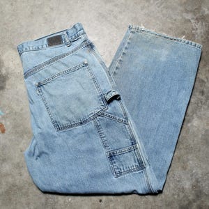 May include: Light blue denim work trousers with multiple pockets and a hammer loop. The jeans have a faded appearance and a leather patch on the back waistband. The trousers are folded on a concrete surface.