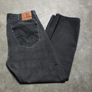 May include: Dark gray denim jeans with a classic five-pocket design. The jeans feature a leather patch and a red tab on the back pocket. The jeans are folded and laid flat on a concrete surface.