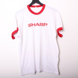 May include: White t-shirt with red trim around the collar and sleeves. The shirt has the word "SHARP" printed in red on the front.
