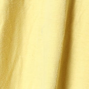 Puede incluir: Primer plano de una tela amarilla p&aacute;lido texturizada. El material tiene una superficie ligeramente irregular con sutiles variaciones de tono, creando una apariencia suave y acogedora. La tela se pliega en pliegues suaves, lo que sugiere un textil ligero.
