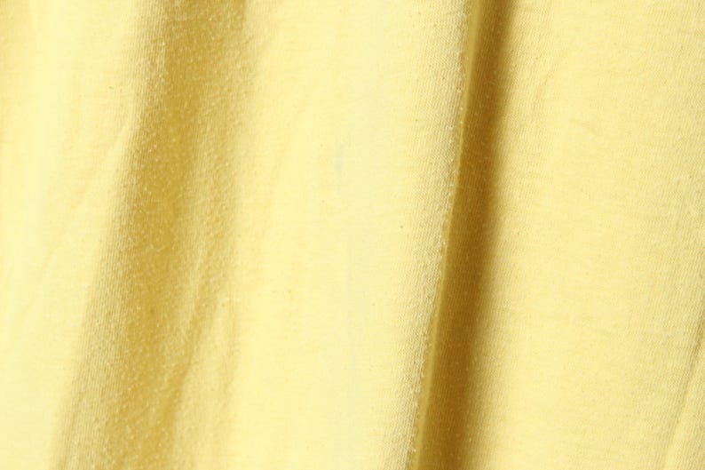 Puede incluir: Primer plano de una tela amarilla p&aacute;lido texturizada. El material tiene una superficie ligeramente irregular con sutiles variaciones de tono, creando una apariencia suave y acogedora. La tela se pliega en pliegues suaves, lo que sugiere un textil ligero.