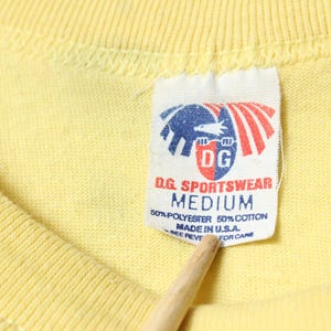 Puede incluir: Primer plano de una camiseta amarilla con una etiqueta. La etiqueta presenta un logotipo con un &aacute;guila azul y rayas rojas, y el texto "D.G. SPORTSWEAR MEDIUM". La camiseta est&aacute; hecha de 50% poli&eacute;ster y 50% algod&oacute;n, fabricada en EE. UU.
