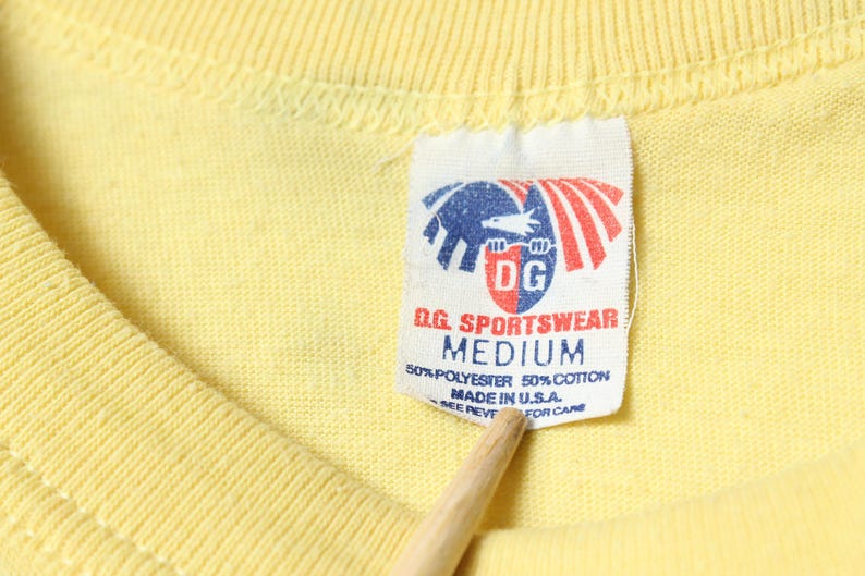 Puede incluir: Primer plano de una camiseta amarilla con una etiqueta. La etiqueta presenta un logotipo con un &aacute;guila azul y rayas rojas, y el texto "D.G. SPORTSWEAR MEDIUM". La camiseta est&aacute; hecha de 50% poli&eacute;ster y 50% algod&oacute;n, fabricada en EE. UU.