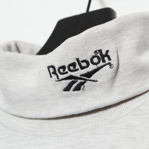 Op de afbeelding: Een grijze trui met een hoge hals en een zwart geborduurd Reebok-logo op de voorkant.