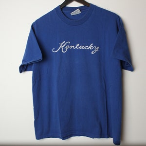 May include: A blue t-shirt with a white embroidered cursive word "Kentucky" on the front.