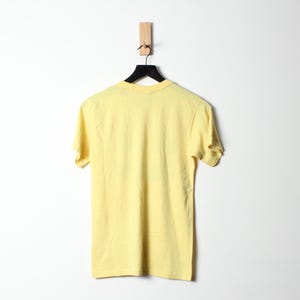 Puede incluir: Una camiseta amarilla p&aacute;lido cuelga de una percha negra sobre un fondo blanco. La camiseta de manga corta est&aacute; hecha de un material suave y se muestra de espaldas al espectador. La percha est&aacute; suspendida de una clavija de madera.