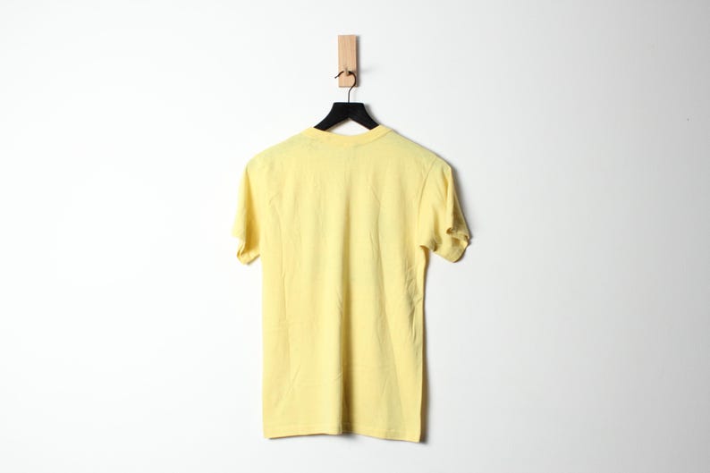 Puede incluir: Una camiseta amarilla p&aacute;lido cuelga de una percha negra sobre un fondo blanco. La camiseta de manga corta est&aacute; hecha de un material suave y se muestra de espaldas al espectador. La percha est&aacute; suspendida de una clavija de madera.