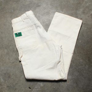 May include: Cream-coloured work trousers with a green and white logo patch on the back pocket. The trousers are folded and appear to be made of a durable material, with multiple pockets and a classic workwear design.