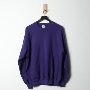 Op de afbeelding: Een effen paarse sweatshirt hangt aan een zwarte hanger tegen een witte muur. De crew neck sweatshirt heeft lange mouwen en een geribbelde zoom en manchetten. Een klein label is zichtbaar op de borst.