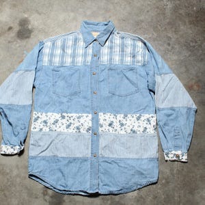 May include: Light blue denim shirt with a patchwork design. The shirt has a collared neckline, button-down front, and long sleeves. The patchwork includes plaid, striped, and floral patterns. The cuffs and bottom hem have a floral pattern.