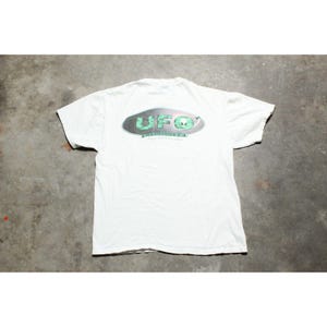 May include: White t-shirt with a graphic on the back featuring the word "UFO" in a metallic oval design with a green alien head. The shirt is made of a soft, lightweight fabric.