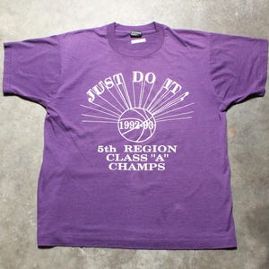 May include: Purple t-shirt with white text reading "JUST DO IT!" above a basketball graphic and "1992-93". Below the graphic, it says "5th REGION CLASS 'A' CHAMPS". The shirt has short sleeves and a crew neck.