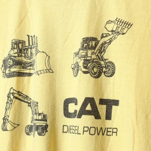 Puede incluir: Camiseta amarilla clara con dibujos en negro de veh&iacute;culos de construcci&oacute;n: una excavadora, una cargadora frontal y una excavadora. El texto "CAT Diesel Power" est&aacute; impreso en letras negras debajo.