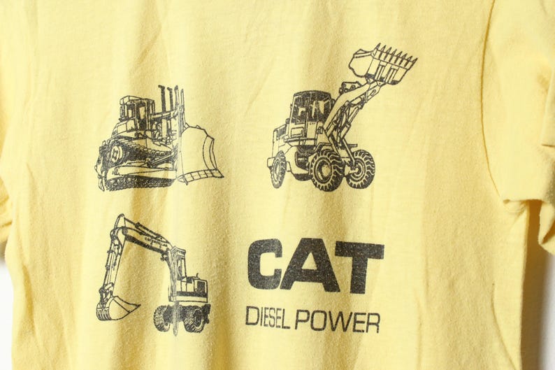 Puede incluir: Camiseta amarilla clara con dibujos en negro de veh&iacute;culos de construcci&oacute;n: una excavadora, una cargadora frontal y una excavadora. El texto "CAT Diesel Power" est&aacute; impreso en letras negras debajo.
