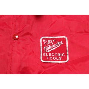 以下が含まれることがあります： 鮮やかな赤色のナイロンジャケット。HEAVY DUTY Milwaukee ELECTRIC TOOLSと白の刺繍文字が入った正方形のパッチが付いています。ジャケットの左側には、赤いスナップボタンが1つ見えています。