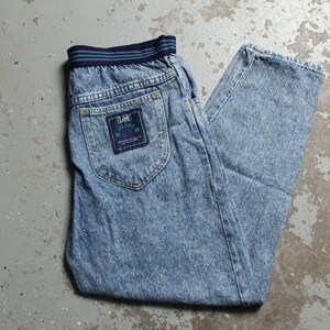 Könnte beinhalten: Hellblaue Jeans aus Denim mit einem elastischen Bund in Lila und Grün. Die Jeans haben eine Gesäßtasche mit einem blauen Etikett, auf dem "Lee Jeans" steht, und ein kleines Logo mit einem Kreis und den Buchstaben "S.L.O."