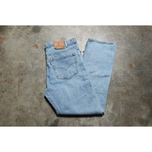 May include: Light blue denim jeans with a classic five-pocket design. The jeans have a brown leather patch on the back waistband and a faded wash. The jeans are folded and laid flat.