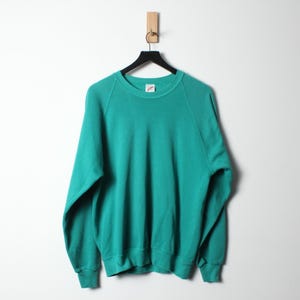 Op de afbeelding: Een turquoise sweatshirt hangt aan een zwarte hanger tegen een witte achtergrond. De crew neck sweatshirt heeft lange mouwen en geribde manchetten en zoom. Het merklabel is zichtbaar bij de halslijn.