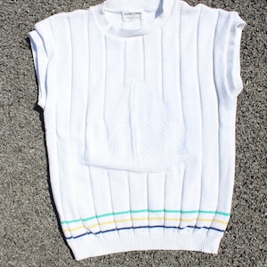May include: White sleeveless knitted sweater with a striped bottom in blue, green, and yellow. The sweater has a small pocket on the front and a label that reads "Alfred Dunner".